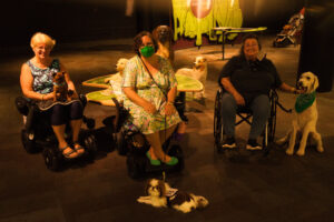 Three happy service dog teams in three wheelchairs face the camera in a museum's dog-themed exhibit.