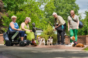 In a lush park, four service dog teams smile at the camera while posing around bronze statues of playing children. The teams have two power wheelchairs and four small dogs.
