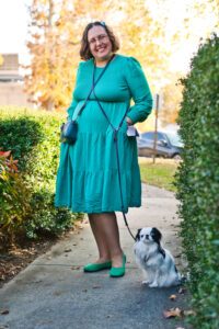 A woman in a bright green dress stands next to a small Japanese Chin service dog sitting on the ground.